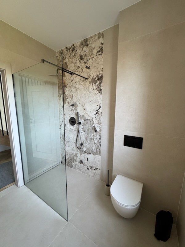 Nowoczesna łazienka z kabiną typu walk-in, wykończona płytkami imitującymi marmur i beżowymi. Minimalistyczna toaleta podwieszana i czarne akcenty.