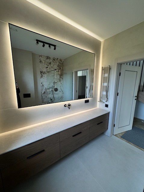 Nowoczesna łazienka z podświetlanym lustrem, drewnianą szafką pod umywalką i grzejnikiem drabinkowym. W lustrze odbija się kabina prysznicowa z dekoracyjną ścianą.