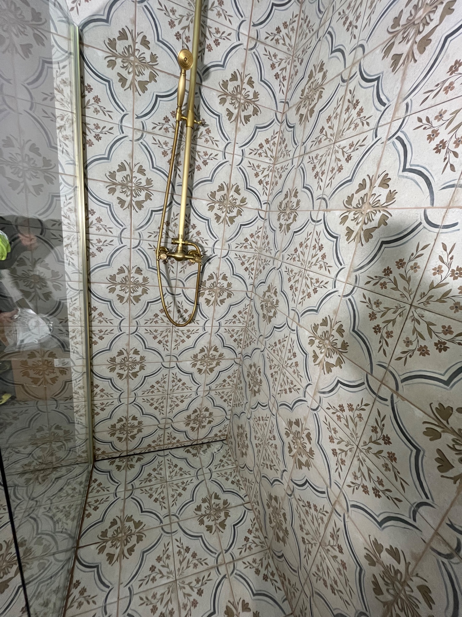 Wykończona łazienka z kabiną prysznicową i złotą armaturą. Płytki w kwiatowy wzór na ścianach i podłodze. Widok z perspektywy narożnika.