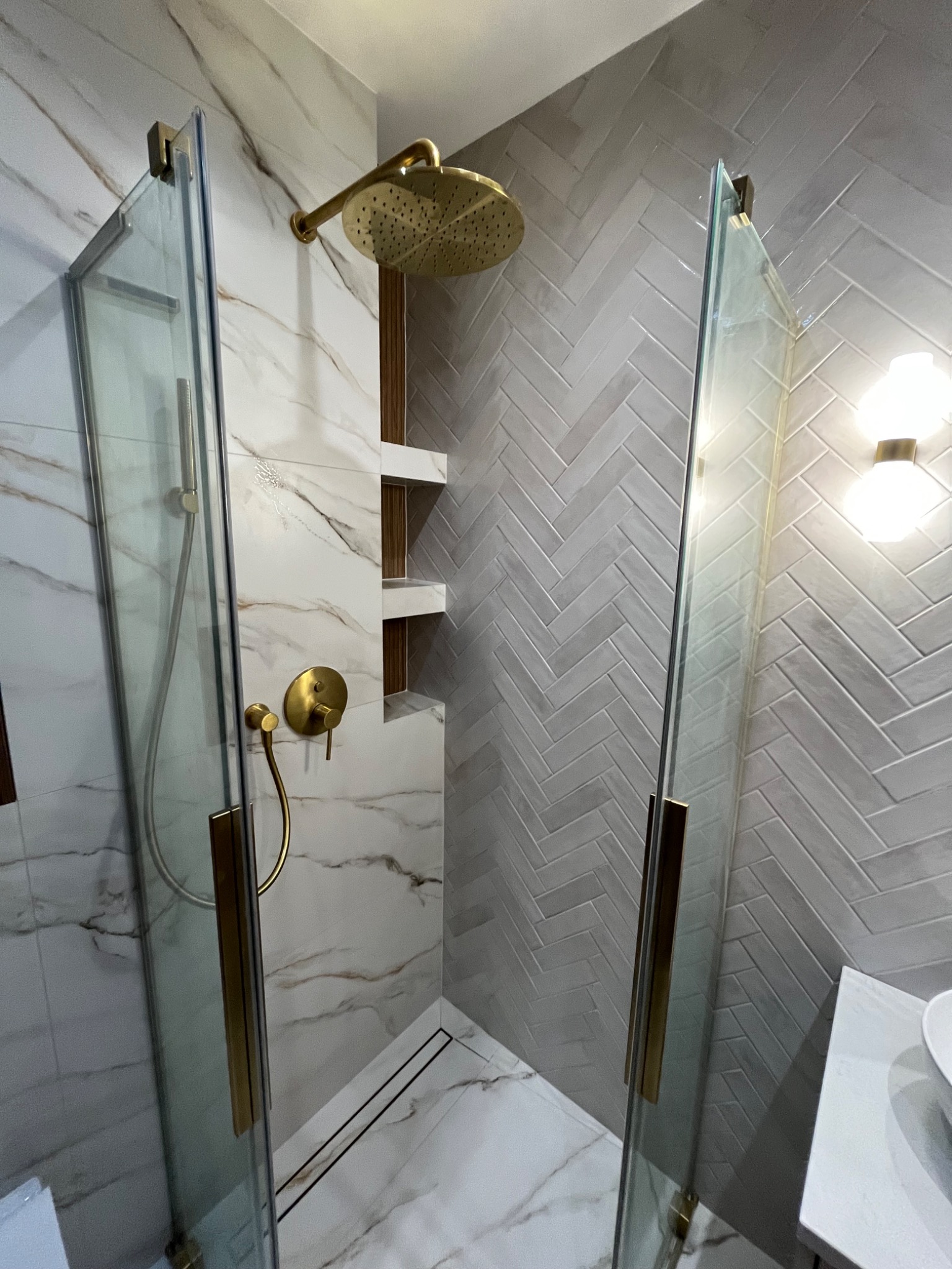 Nowoczesna kabina prysznicowa z marmurowymi płytkami i złotą armaturą. Ściana wyłożona płytkami w jodełkę. Minimalistyczny design z otwartą przestrzenią.