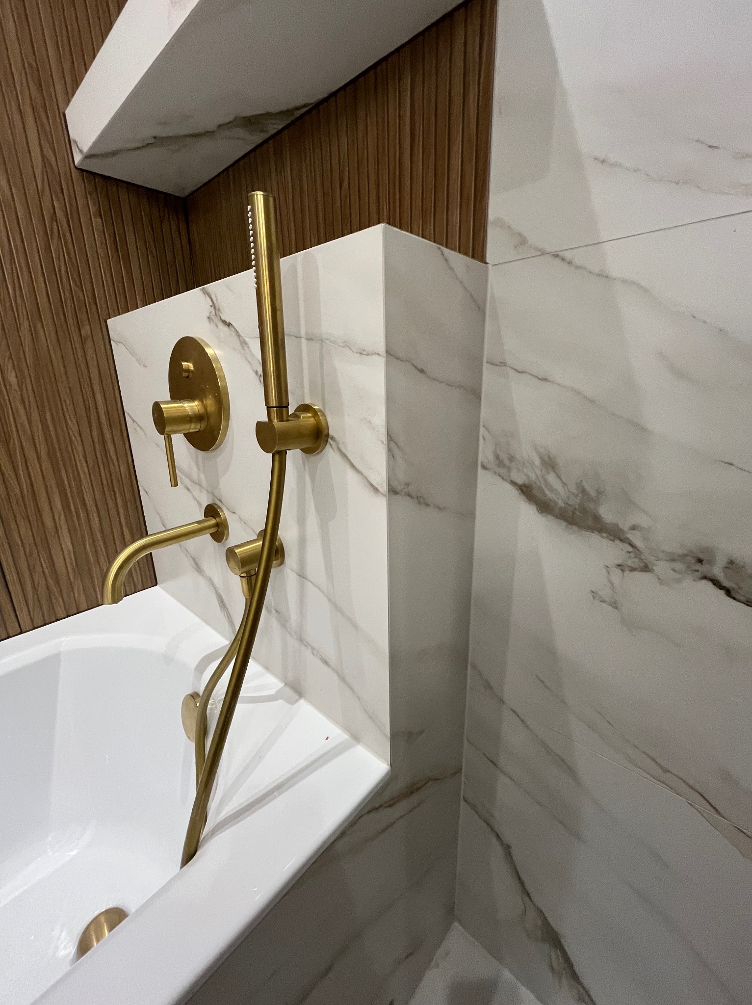 Biała wanna z marmurową obudową i złotą armaturą, w łazience z drewnianymi i marmurowymi płytkami. Ujęcie z bliska, prezentujące detale wykończenia.