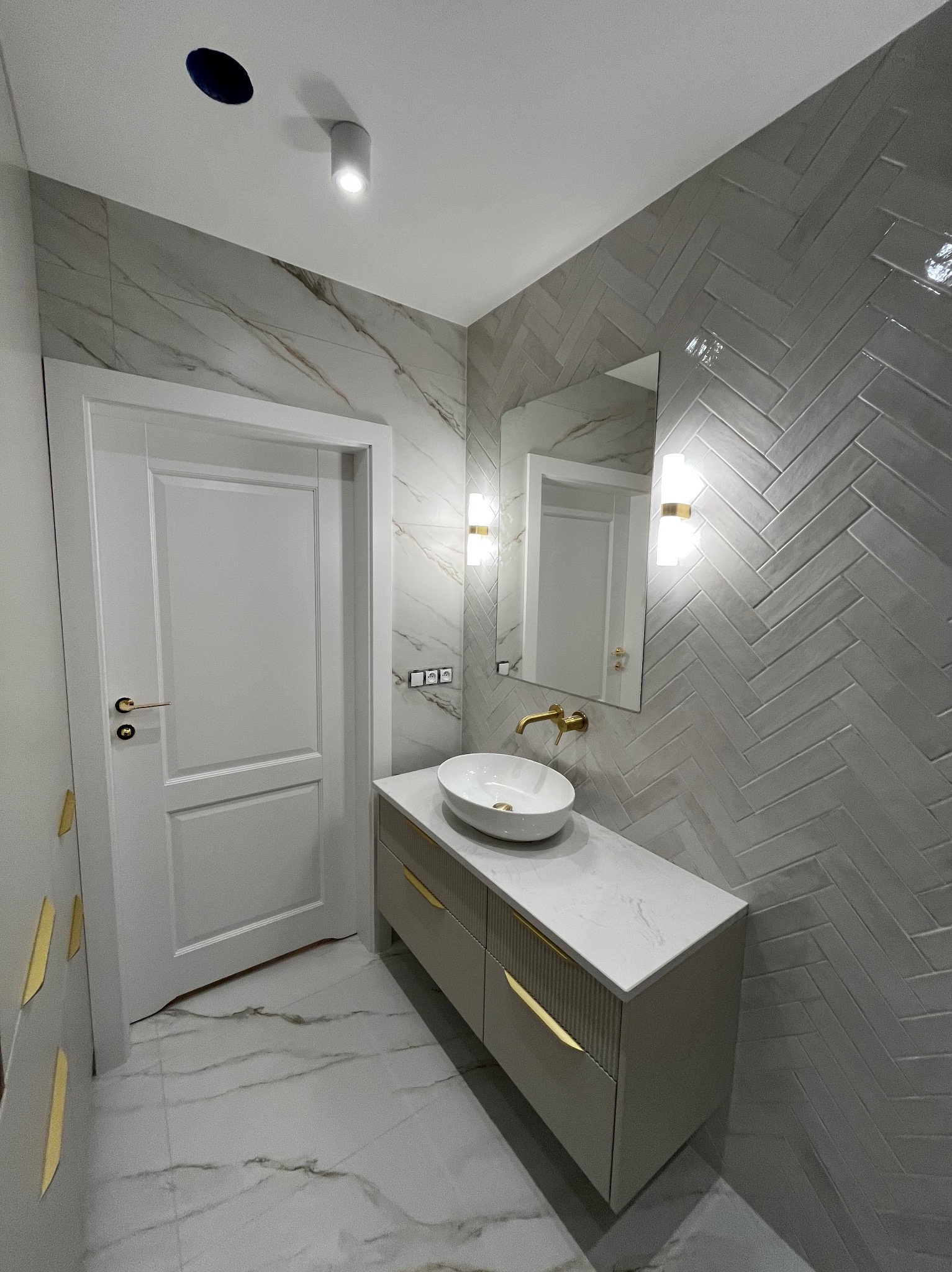 Elegancka łazienka z umywalką nablatową, złotą armaturą i szarą glazurą ułożoną w jodełkę. Białe drzwi i marmurowe płytki podłogowe dopełniają nowoczesny wystrój.