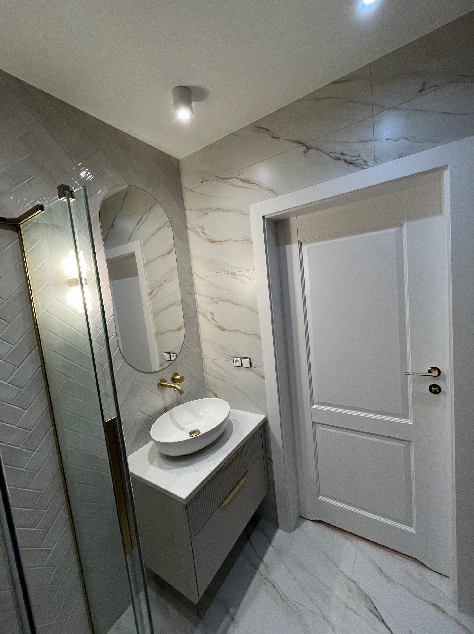 Nowoczesna łazienka z owalnym lustrem, białą umywalką nablatową i kabiną prysznicową z złotymi detalami. Marmurowe płytki na ścianach i podłodze.