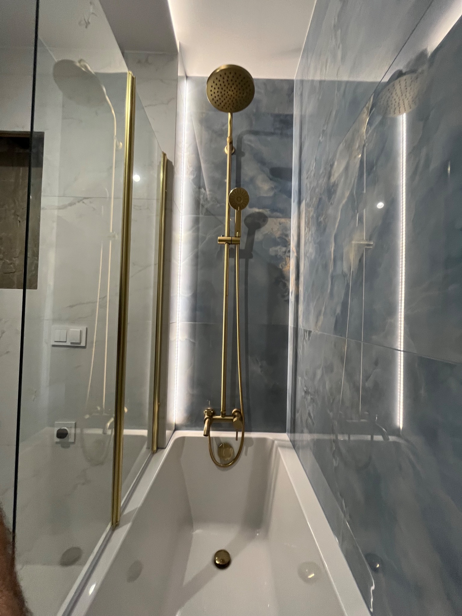 Nowoczesna łazienka z wanną, złotą armaturą i marmurowymi płytkami w odcieniach błękitu. Oświetlenie LED podkreśla elegancki design. Widok na kabinę prysznicową.