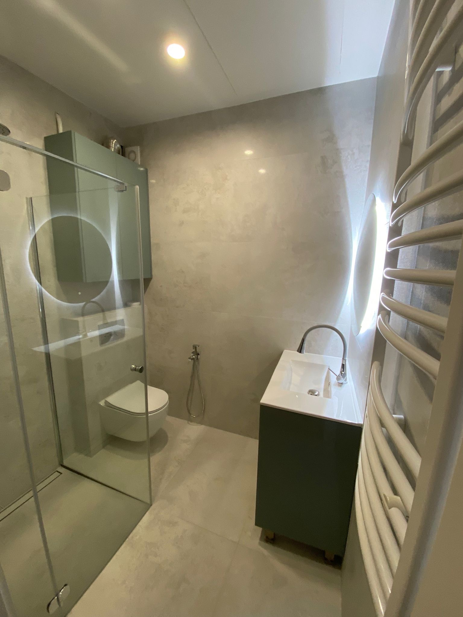 Nowoczesna łazienka z kabiną prysznicową typu walk-in, umywalką na szafce w odcieniach zieleni i podświetlanym lustrem. Minimalistyczny design, jasne płytki i grzejnik drabinkowy.