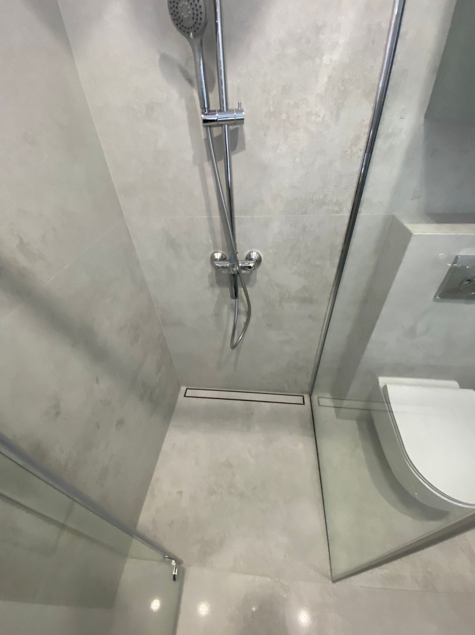 Nowoczesna kabina prysznicowa z odpływem liniowym i szarą, teksturowaną płytką. Minimalistyczny design, chromowana armatura, częściowo widoczna toaleta podtynkowa.