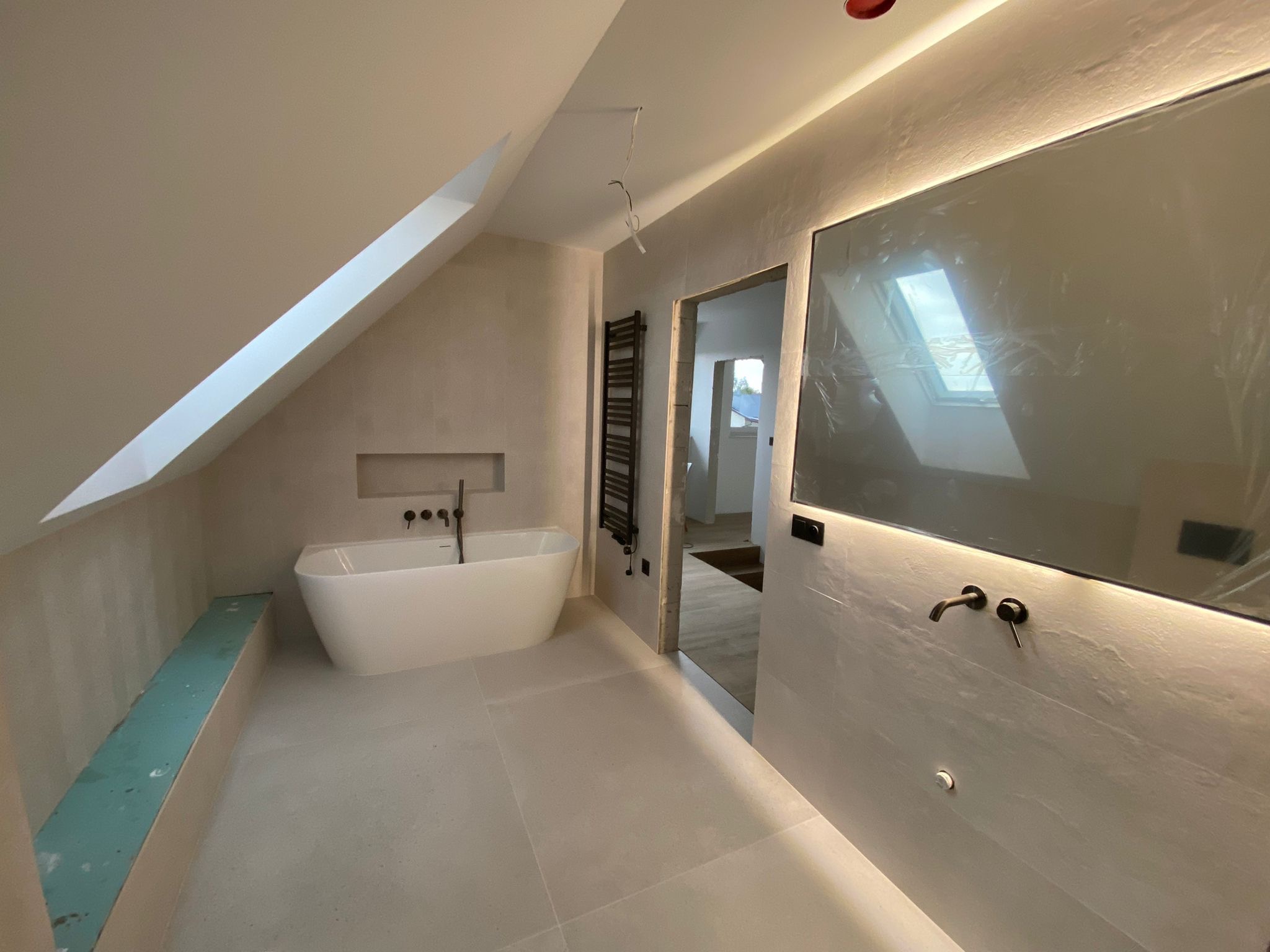 Nowoczesna łazienka na poddaszu z wolnostojącą wanną, podświetlanym lustrem i grzejnikiem drabinkowym. Minimalistyczny design w jasnych barwach.