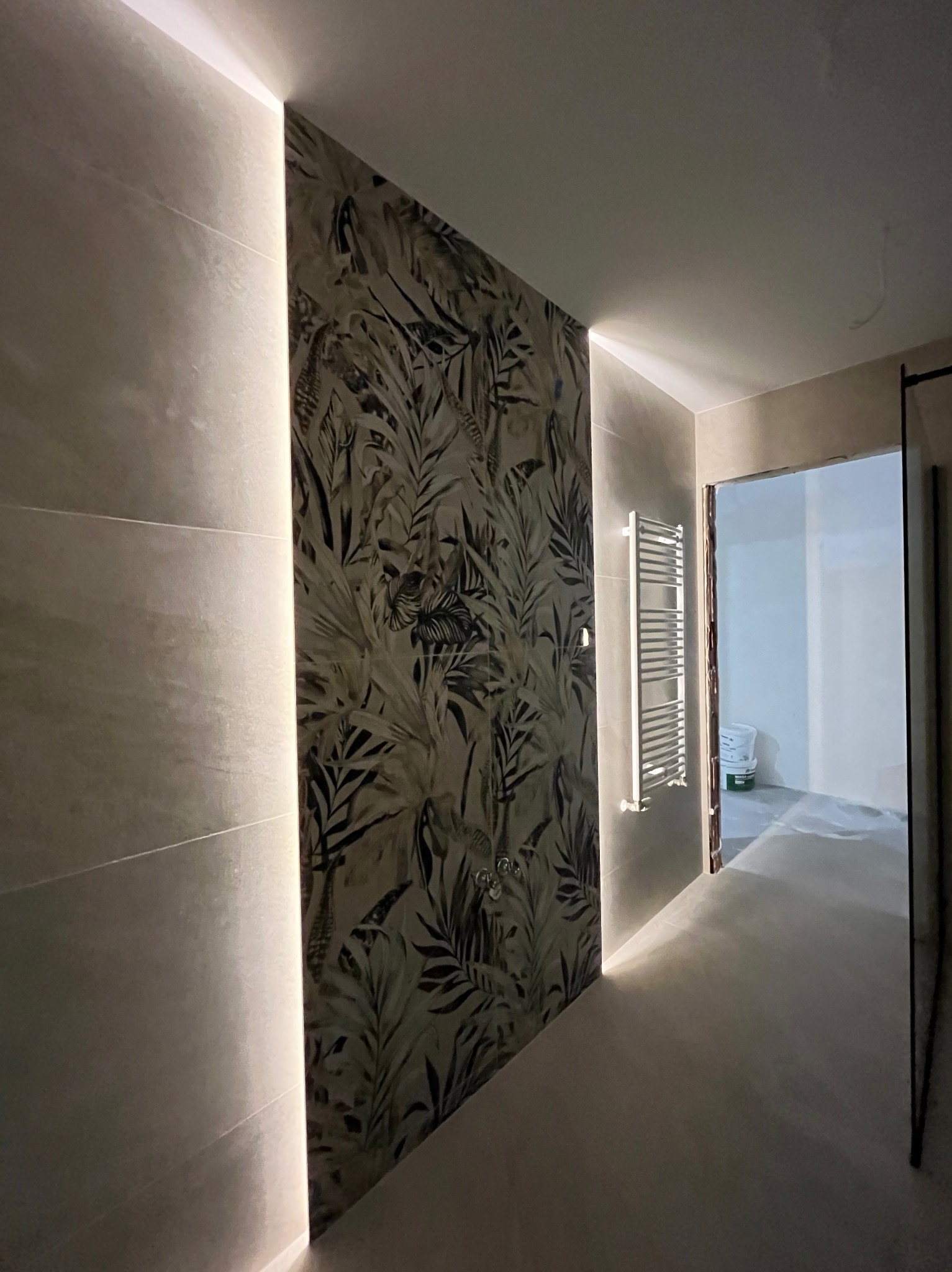 Nowoczesna łazienka z podświetlaną ścianą z motywem roślinnym i grzejnikiem drabinkowym. Widok na dalszą część pomieszczenia w trakcie prac wykończeniowych.