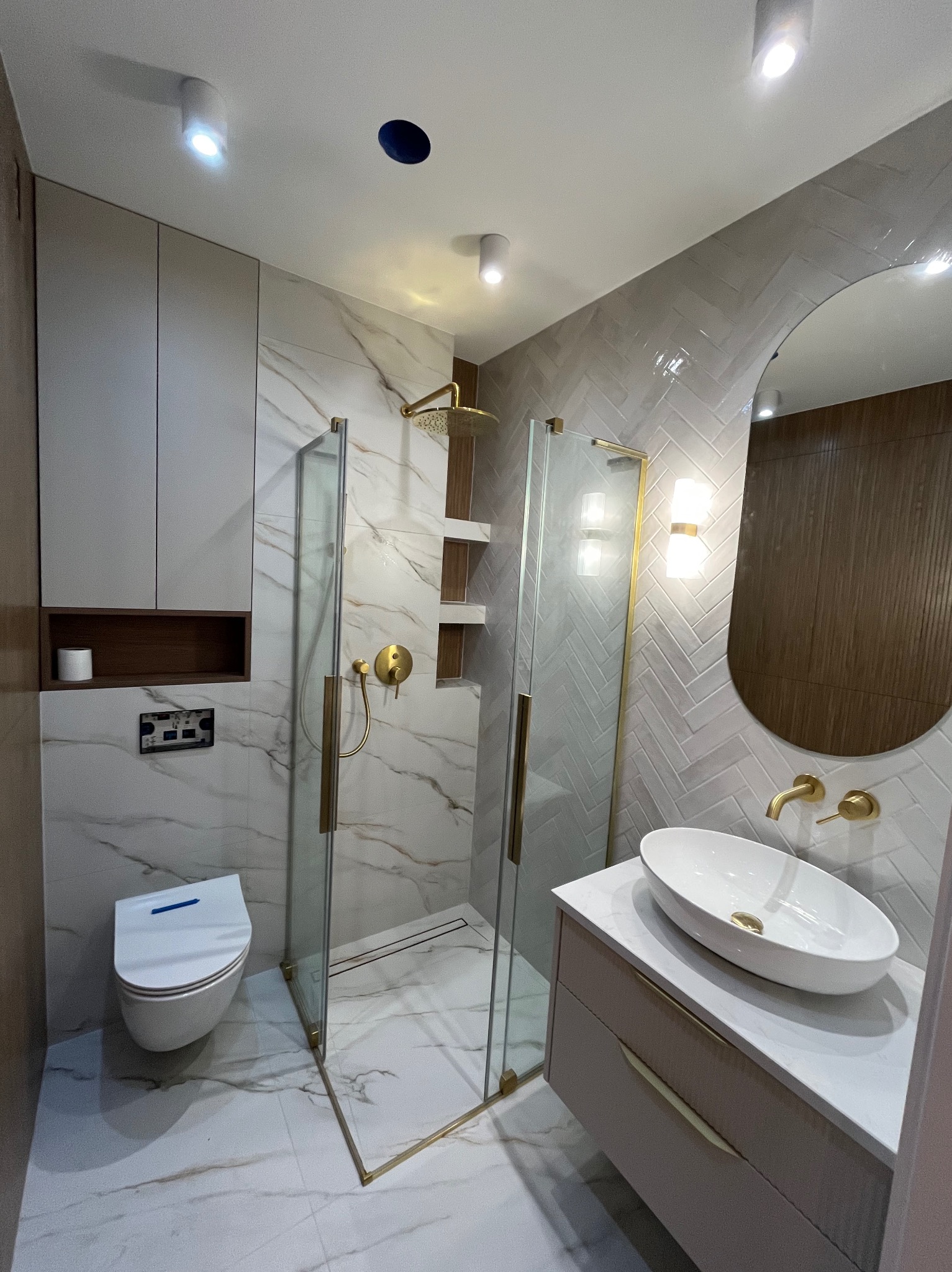 Nowoczesna łazienka po remoncie: kabina prysznicowa ze złotymi detalami, umywalka nablatowa, toaleta podwieszana i płytki imitujące marmur. Oświetlenie punktowe.