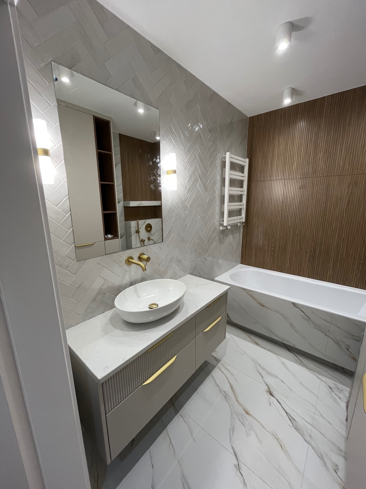 Nowoczesna łazienka z umywalką nablatową, złotą armaturą i płytkami w jodełkę. Uwagę zwraca marmurowa wanna i drewniane panele ścienne.