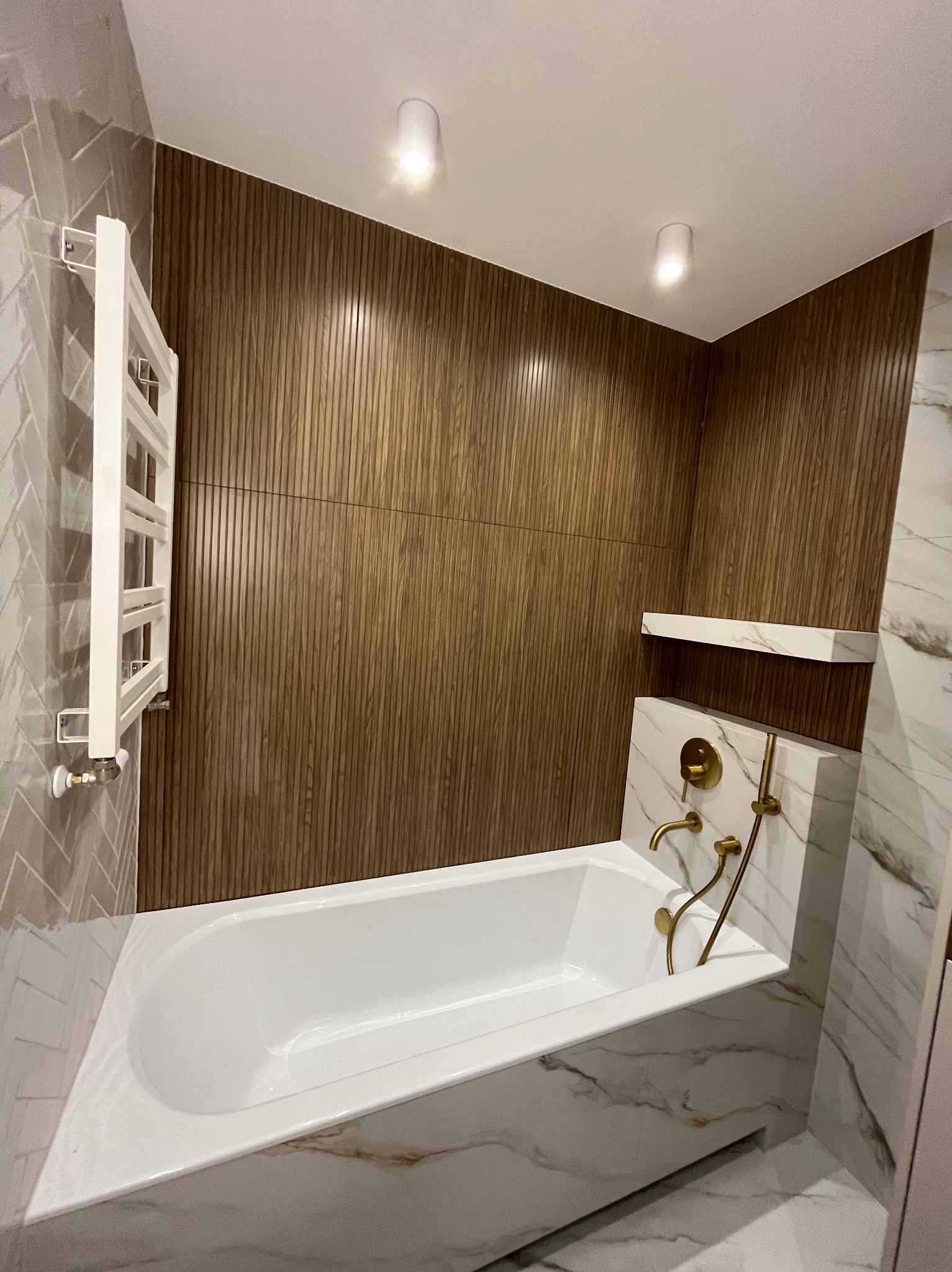 Nowoczesna łazienka z wanną obudowaną marmurem, drewnianą ścianą i złotą armaturą. Minimalistyczny grzejnik drabinkowy i oświetlenie punktowe.