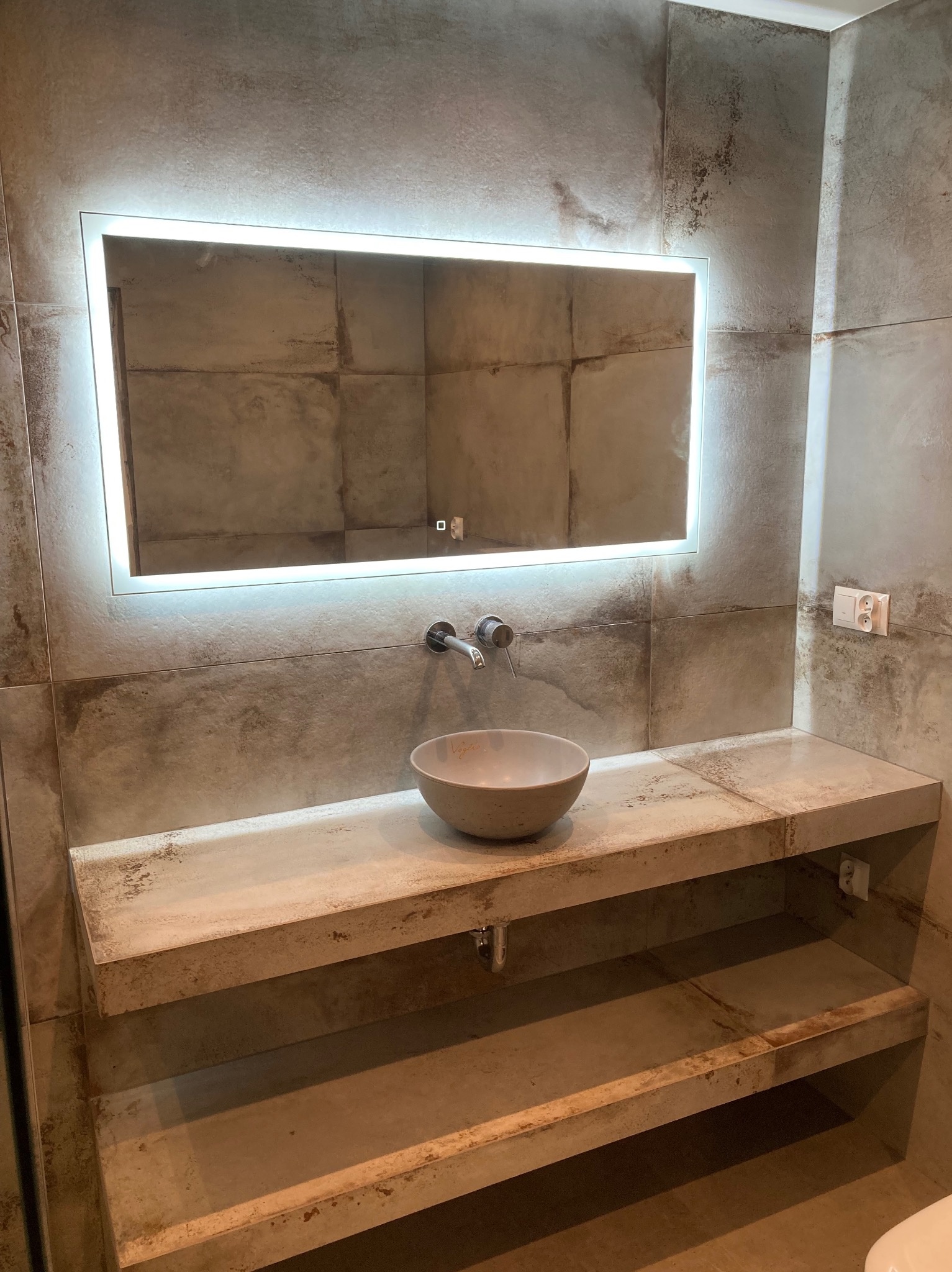 Nowoczesna łazienka z umywalką w kształcie misy, podświetlanym lustrem i półkami z płytek imitujących beton. Minimalistyczny design z widocznymi gniazdkami.