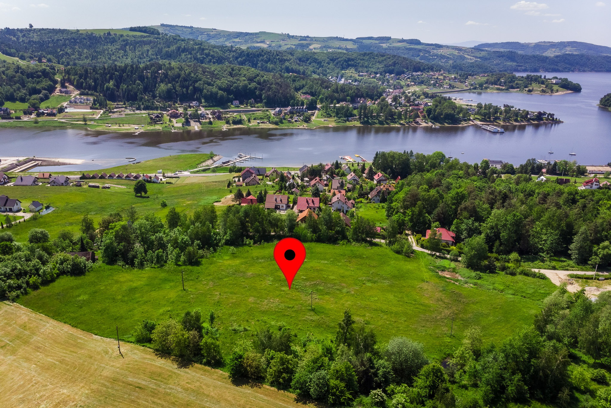 Panoramiczny widok z lotu ptaka na działkę z czerwoną pinezką, w tle jezioro i zabudowa. Słoneczny dzień, bujna zieleń drzew i łąk.