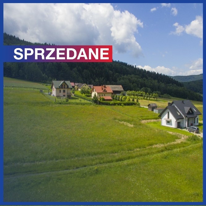 Sprzedany dom na tle zielonego wzgórza i błękitnego nieba z chmurami. Na pierwszym planie łąka, w tle las. Czerwono-biały napis 'SPRZEDANE' w lewym górnym rogu.