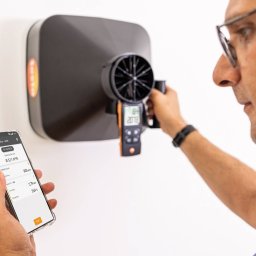 PROinstal - ogrzewanie, chłodnictwo, wentylacja - Mężczyzna mierzy przepływ powietrza z anemostatu za pomocą miernika i aplikacji na smartfonie, kontrolując parametry wentylacji. Widoczny anemostat i wyniki pomiarów.