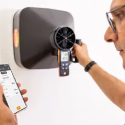 Mężczyzna mierzy przepływ powietrza z anemostatu za pomocą miernika i aplikacji na smartfonie, kontrolując parametry wentylacji. Widoczny anemostat i wyniki pomiarów.