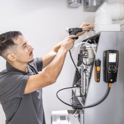 PROinstal - ogrzewanie, chłodnictwo, wentylacja - Mężczyzna w szarej koszulce serwisuje piec gazowy, używając specjalistycznego sprzętu pomiarowego Testo do analizy spalin. Widoczne przewody i elementy pieca.