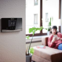 PROinstal - ogrzewanie, chłodnictwo, wentylacja - Nowoczesny panel sterowania ogrzewaniem Vaillant na ścianie w salonie, w tle kobieta na kanapie. Wyświetlacz pokazuje 21 stopni Celsjusza.