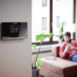 Nowoczesny panel sterowania ogrzewaniem Vaillant na ścianie w salonie, w tle kobieta na kanapie. Wyświetlacz pokazuje 21 stopni Celsjusza.