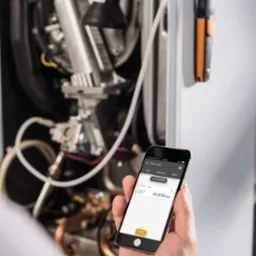 Pomiar ciśnienia gazu w piecu za pomocą smartfona i specjalistycznego urządzenia pomiarowego z pomarańczowymi akcentami, w tle wnętrze kotła.