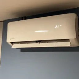 Biały klimatyzator Rotenso z wyświetlaczem 21°C, zamontowany na błękitnej ścianie. Widoczne logo producenta i model. Minimalistyczny design.