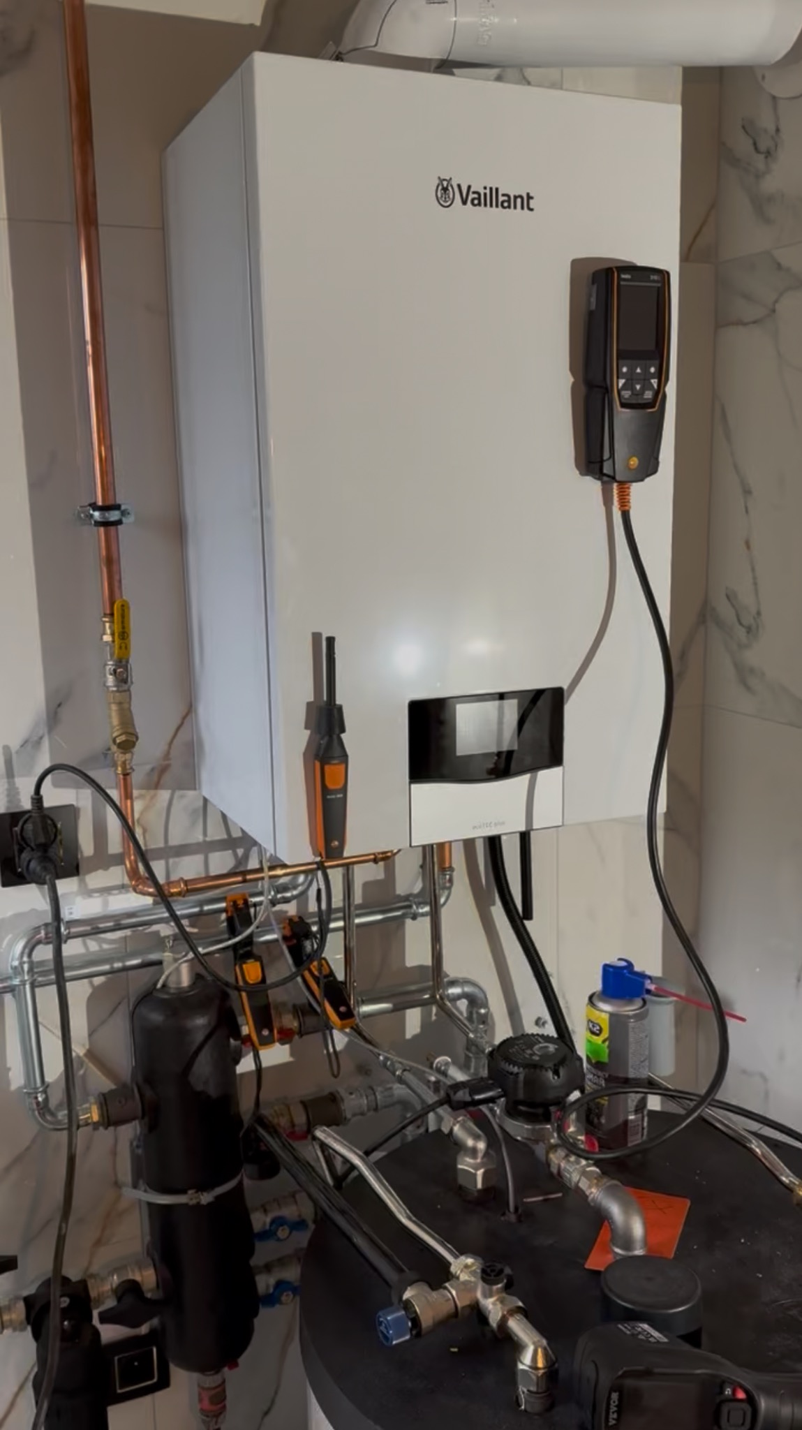 Biały, wiszący piec gazowy Vaillant z podłączonymi rurami miedzianymi i czarnym osprzętem. Widoczne narzędzia pomiarowe i elementy instalacji.