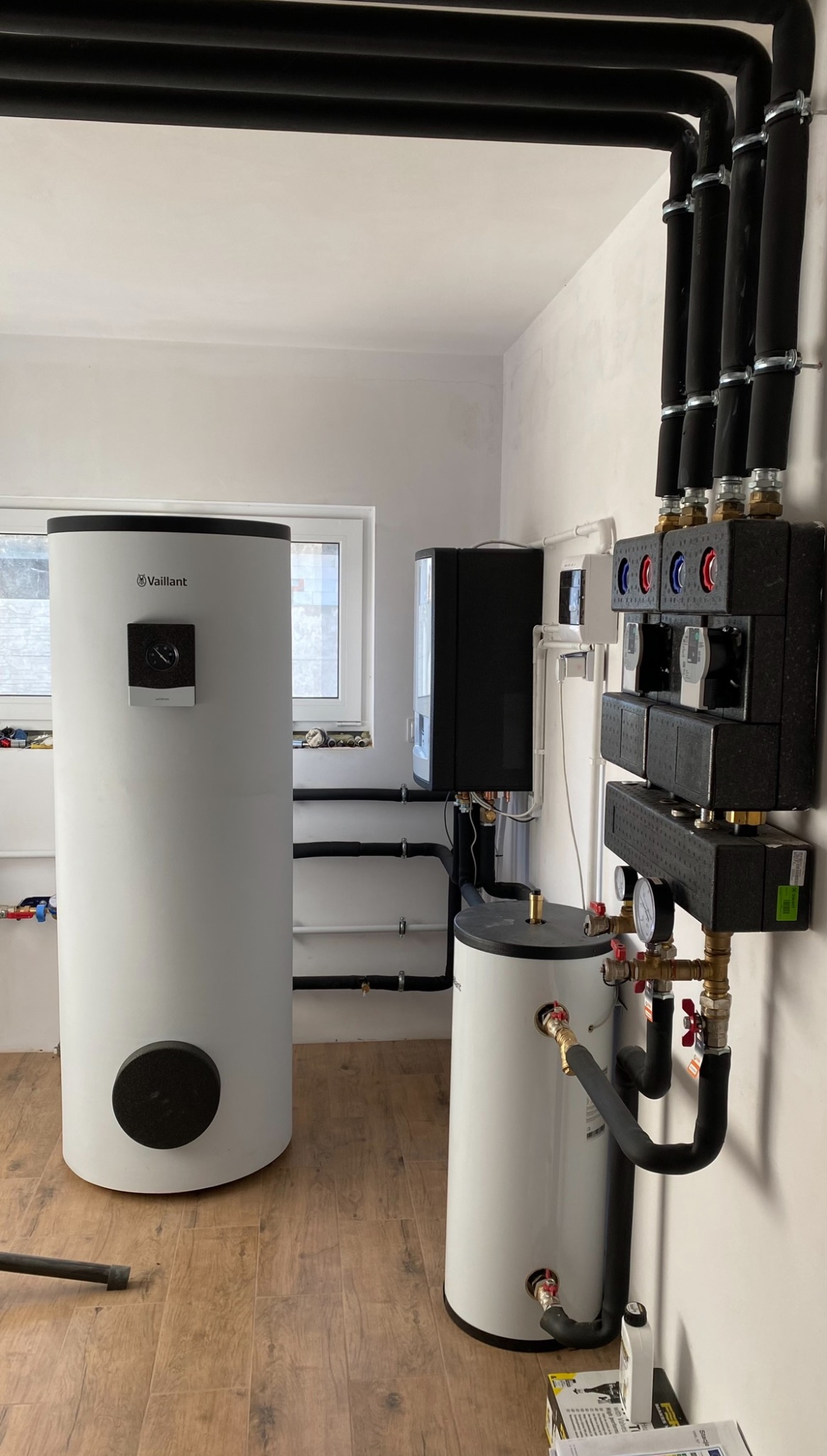 Biały zasobnik ciepłej wody Vaillant z czarnymi rurami w pomieszczeniu z drewnopodobną podłogą. Widoczne zawory i elementy instalacji grzewczej.