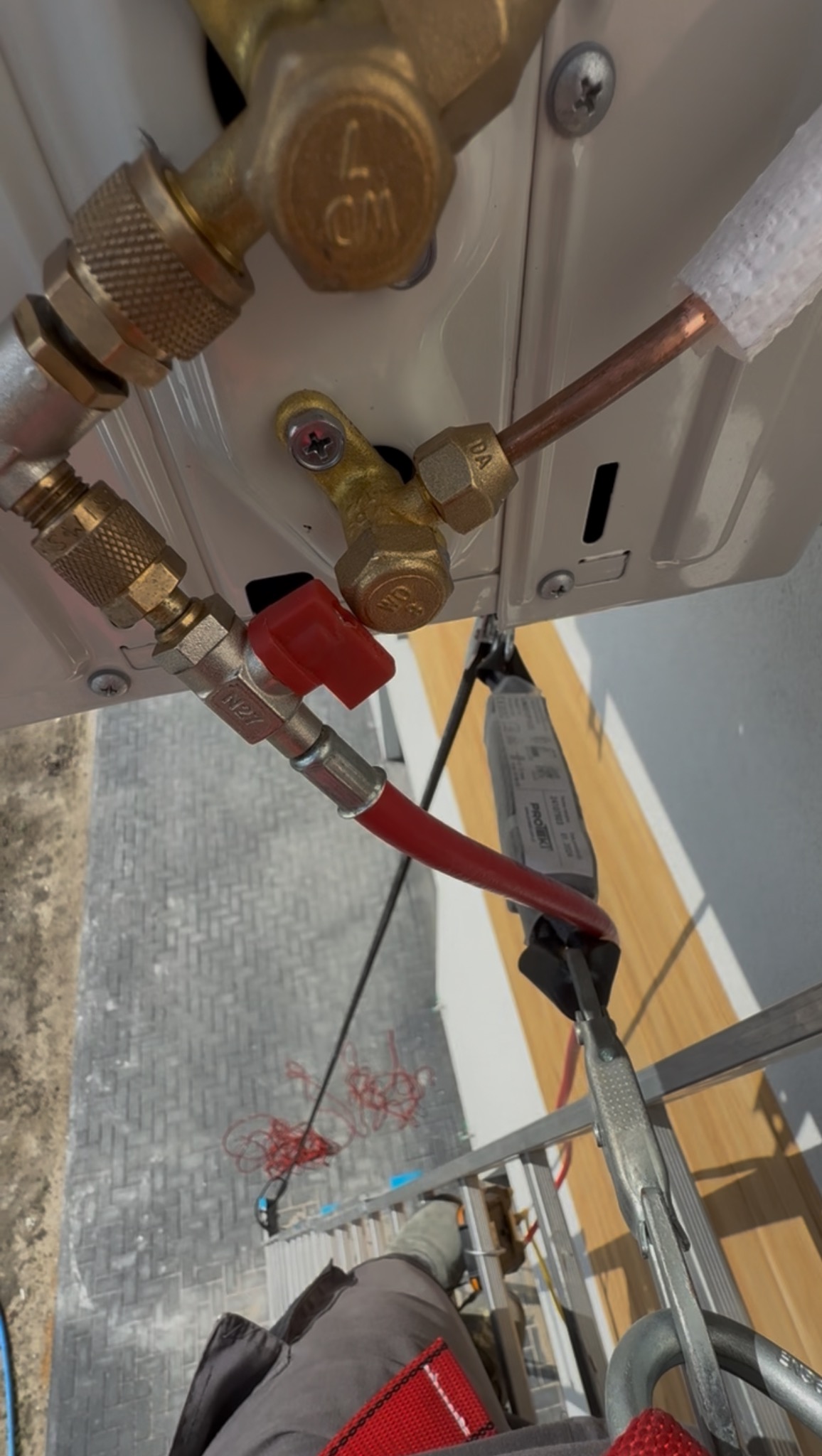 Szczegółowe ujęcie instalacji klimatyzacji z miedzianymi rurami i zaworami, widoczne z perspektywy osoby na drabinie podczas montażu.