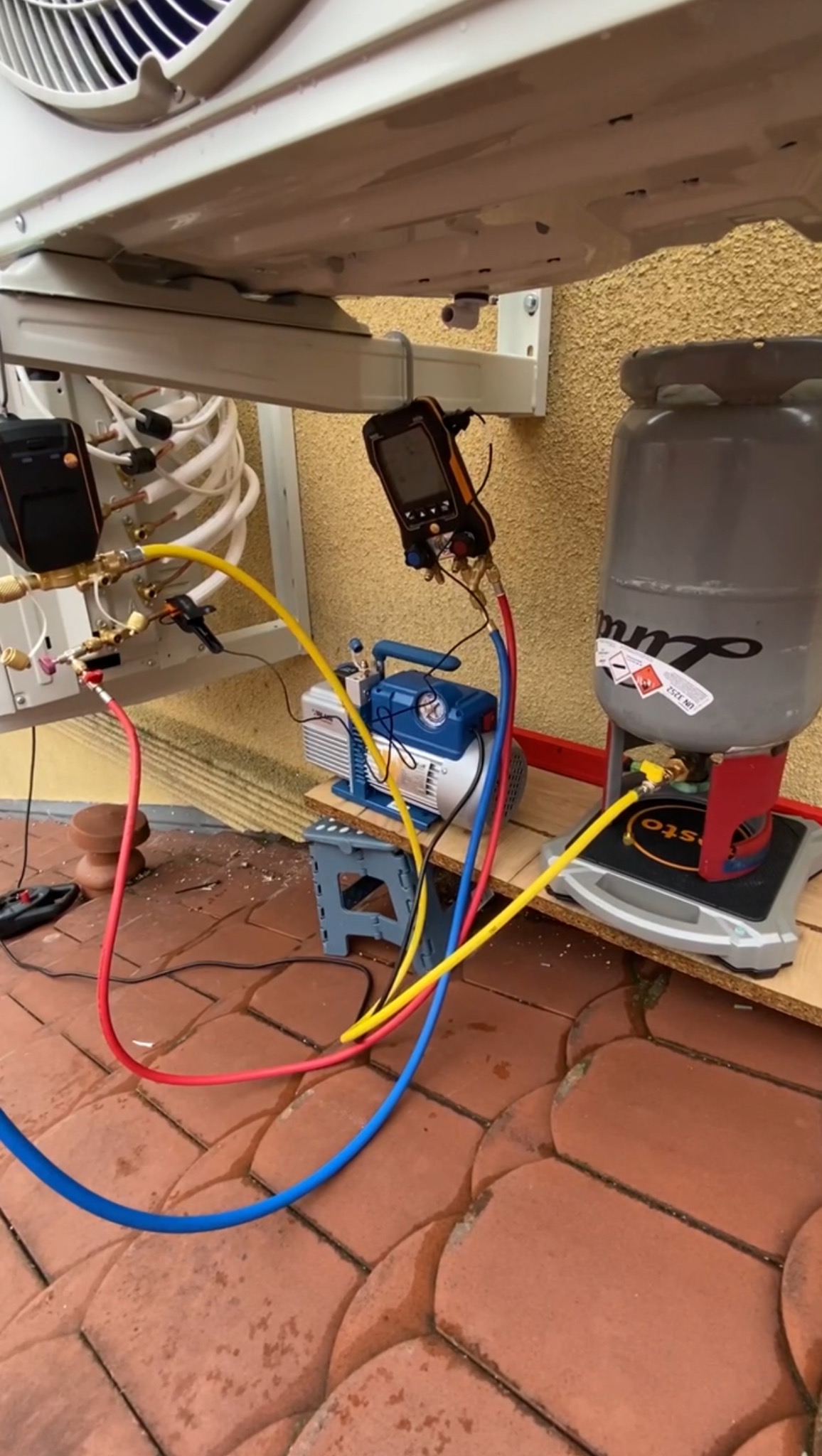 Podłączenie klimatyzacji zewnętrznej: manometry, pompa próżniowa i butla z czynnikiem chłodniczym na tle elewacji budynku i mokrej kostki brukowej.