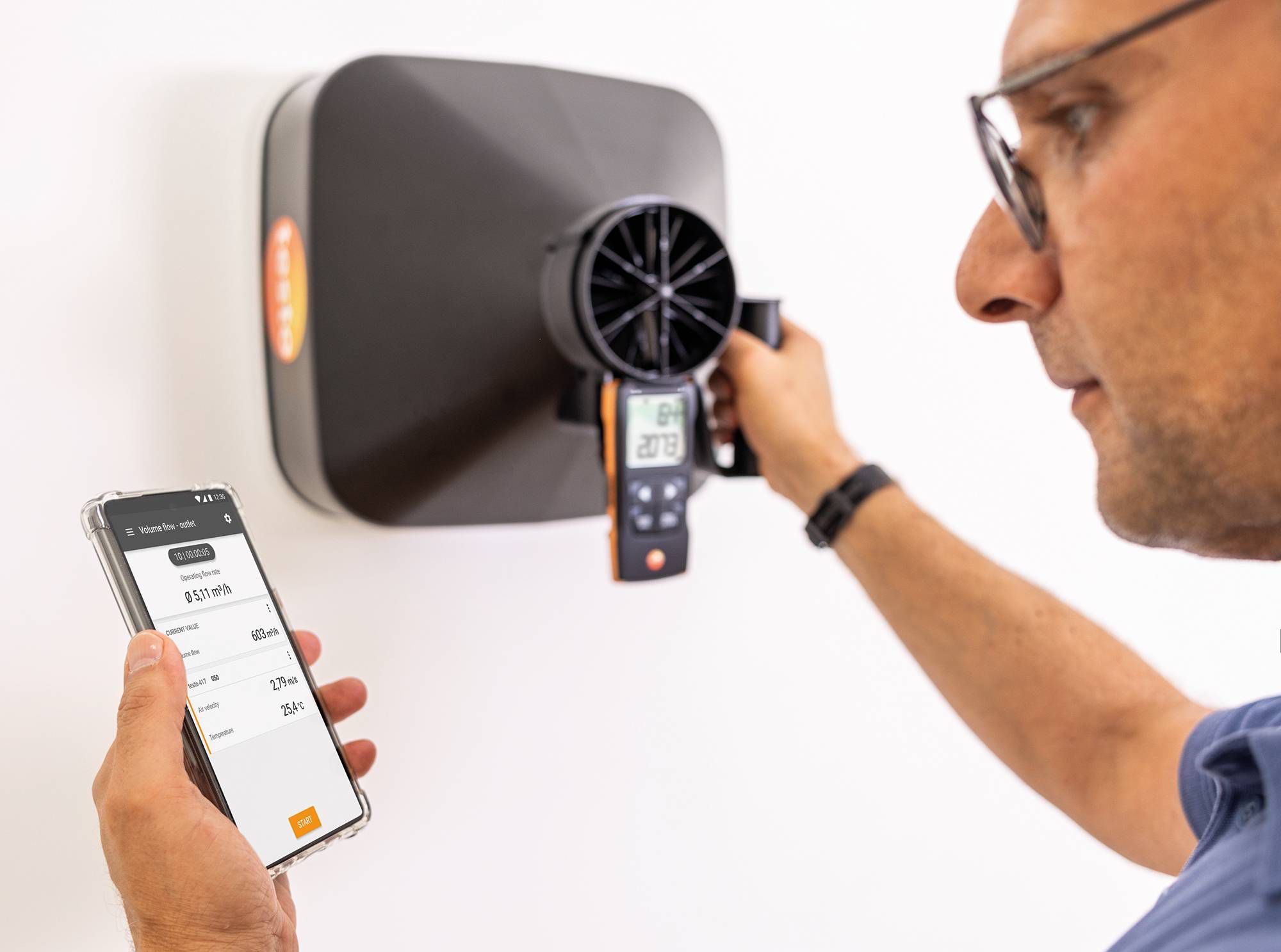 Mężczyzna mierzy przepływ powietrza z anemostatu za pomocą miernika i aplikacji na smartfonie, kontrolując parametry wentylacji. Widoczny anemostat i wyniki pomiarów.