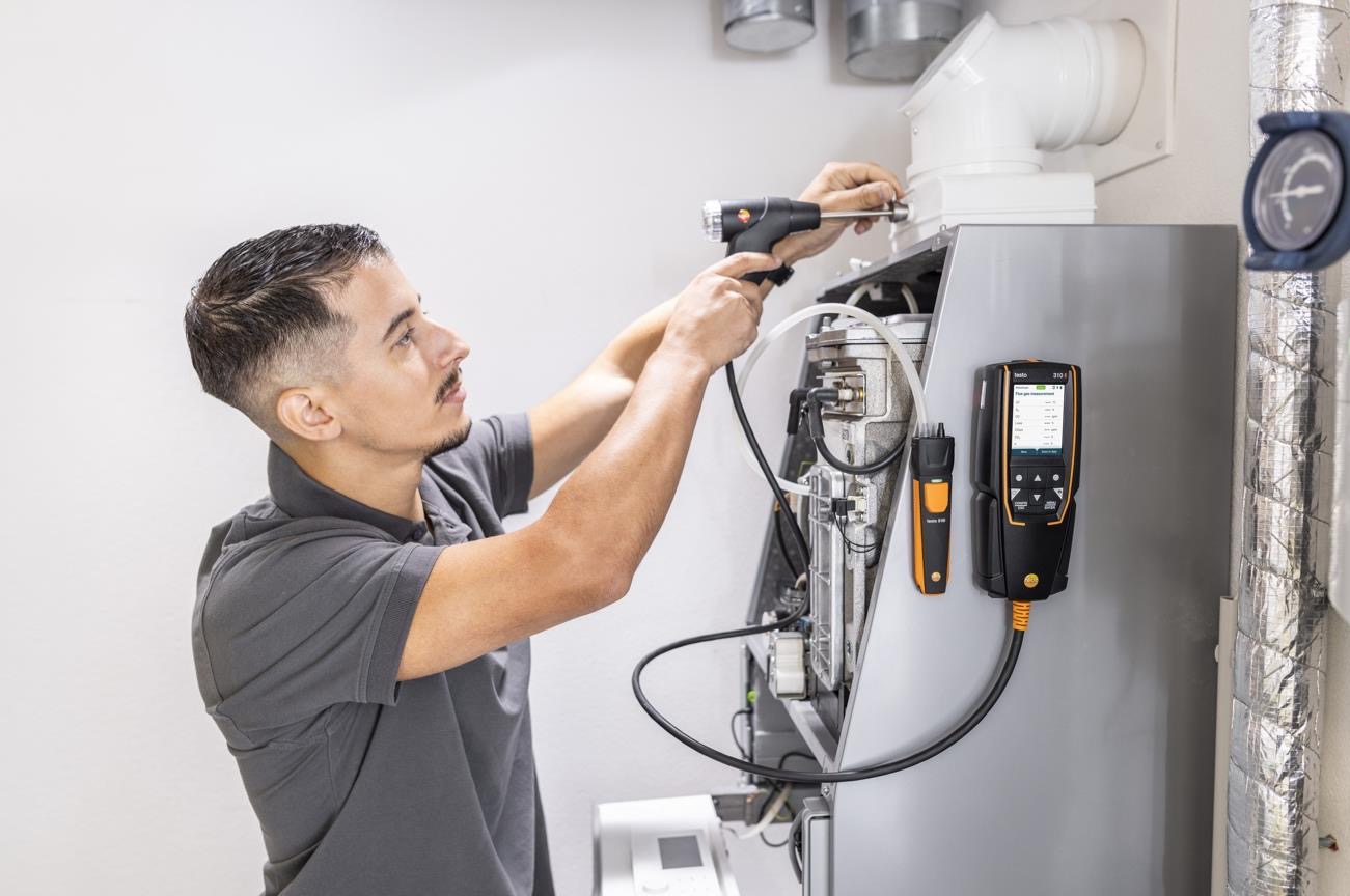 Mężczyzna w szarej koszulce serwisuje piec gazowy, używając specjalistycznego sprzętu pomiarowego Testo do analizy spalin. Widoczne przewody i elementy pieca.