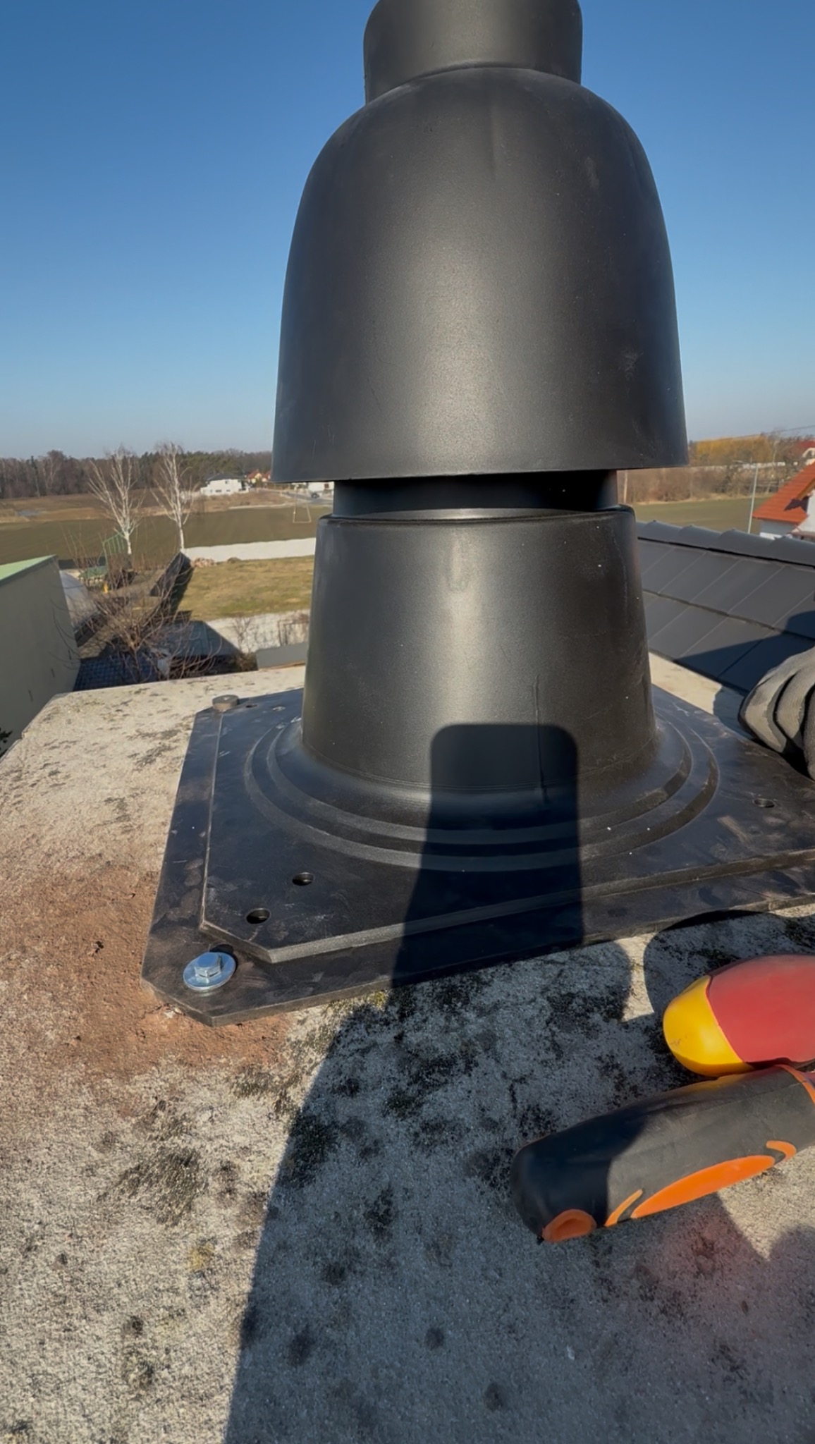 Czarny kominek wentylacyjny na dachu, zamocowany na betonowej podstawie, widoczne narzędzia. Błękitne niebo w tle, częściowo zacieniony przez cień osoby.