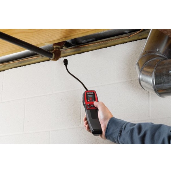 Kontrola szczelności instalacji: detektor gazu w dłoni, rura instalacyjna i komin w tle. Biała ściana z bloków betonowych. Czerwono-czarny detektor.