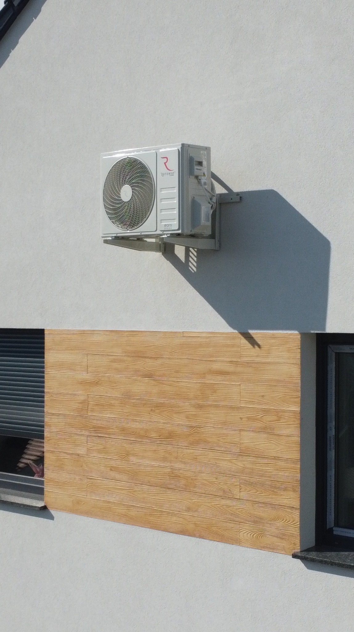 Biała jednostka zewnętrzna klimatyzacji Rotenso zamontowana na ścianie budynku z oknem i imitacją drewnianej okładziny. Widoczny cień urządzenia.