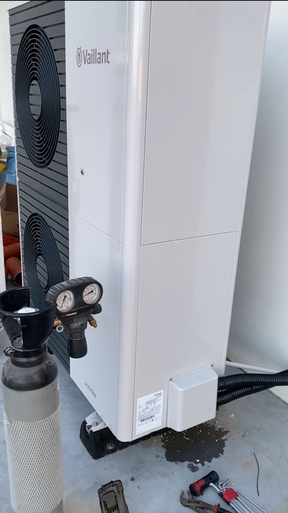 Biała pompa ciepła Vaillant podczas instalacji w Miejskiej Górce, widoczny manometr i butla z czynnikiem chłodniczym oraz narzędzia, na podłodze woda.