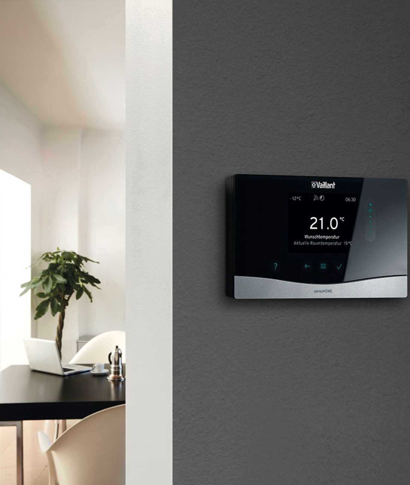 Czarny panel sterowania ogrzewaniem Vaillant sensoHOME na szarej ścianie, ustawiony na 21 stopni Celsjusza. W tle widoczny fragment biura z rośliną i laptopem.