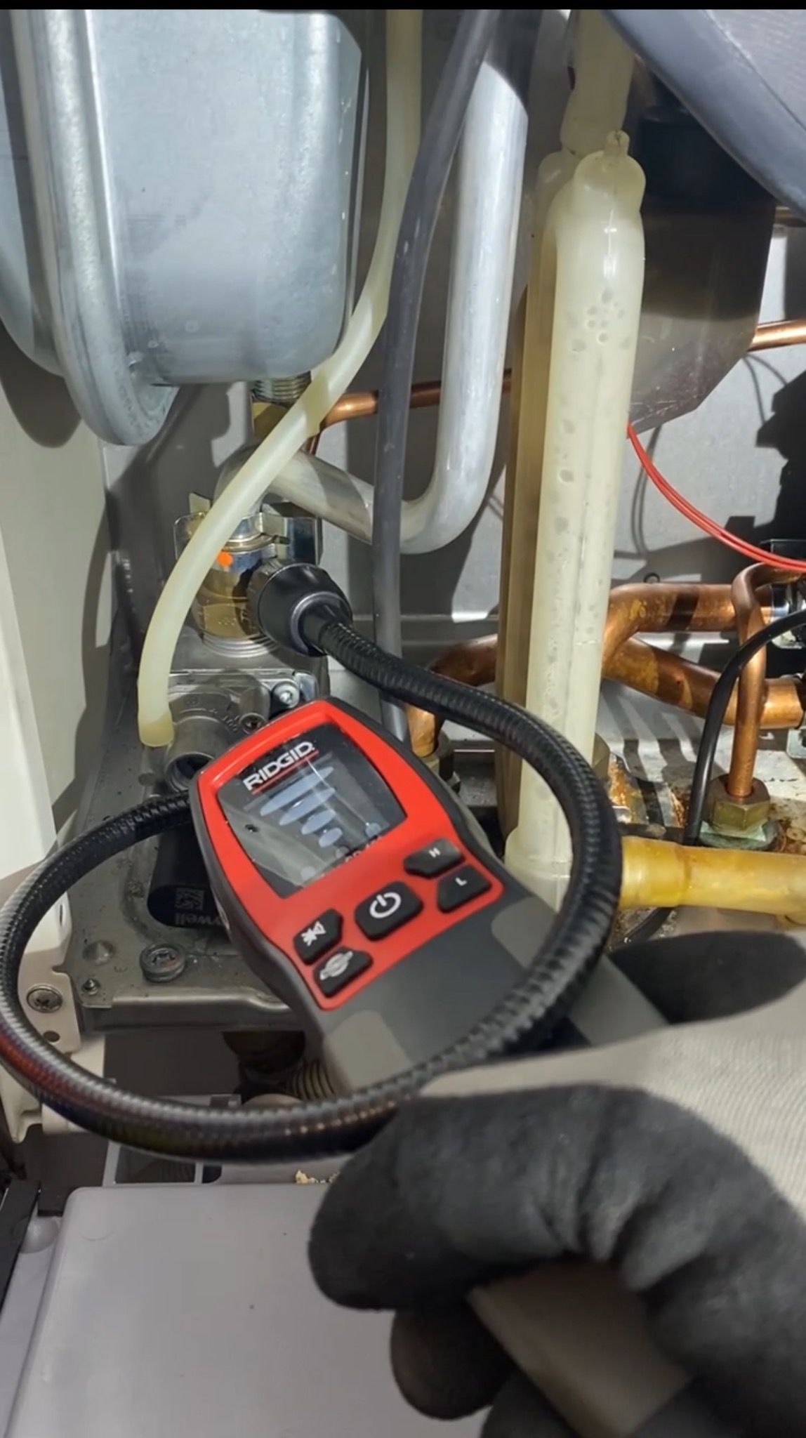 Sprawdzanie szczelności instalacji gazowej za pomocą detektora gazu Ridgid wewnątrz kotła. Widoczne rury miedziane i plastikowe.