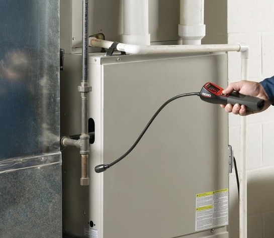 Kontrola szczelności instalacji gazowej za pomocą detektora. Mężczyzna sprawdza urządzeniem rurę przyłączeniową pieca w pomieszczeniu z białymi ścianami.
