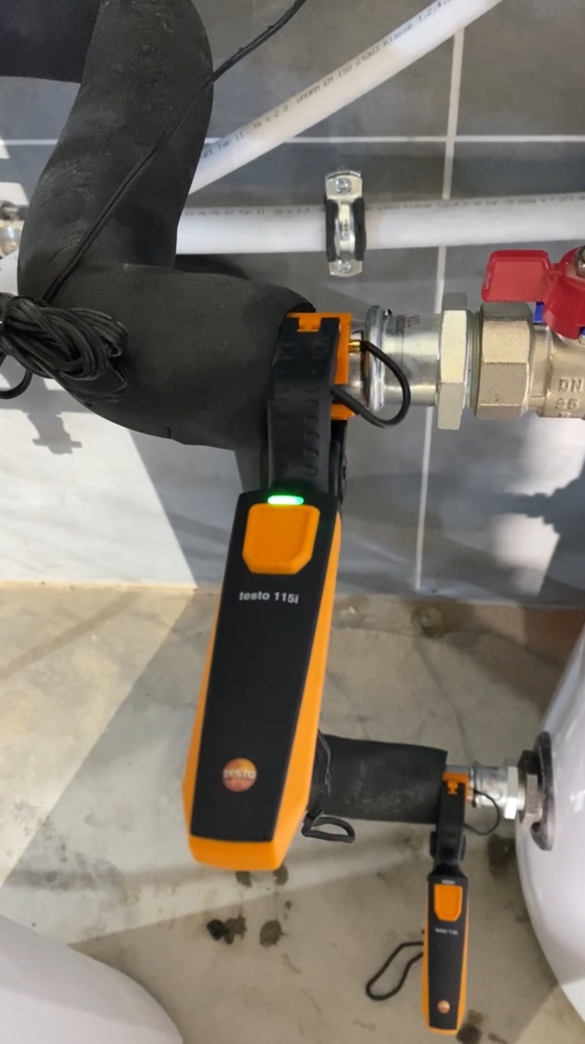 Pomiar temperatury rury grzewczej za pomocą miernika Testo 115i z zieloną diodą, widoczny fragment instalacji i zawór. Szare płytki na podłodze i ścianie.