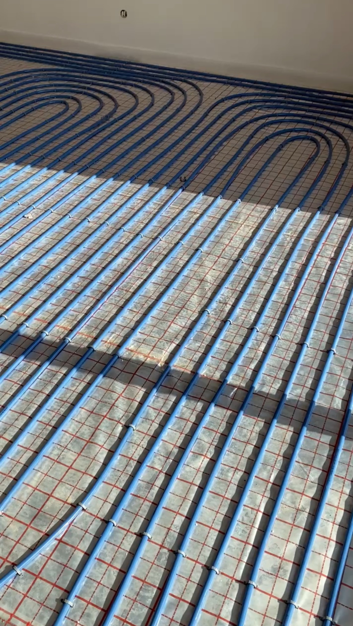 Niskopodłogowe ogrzewanie: błękitne rury ułożone w pętle na folii z czerwoną kratką, przygotowane do zalania posadzką. Widoczne mocowania rur. Częściowe oświetlenie słoneczne.