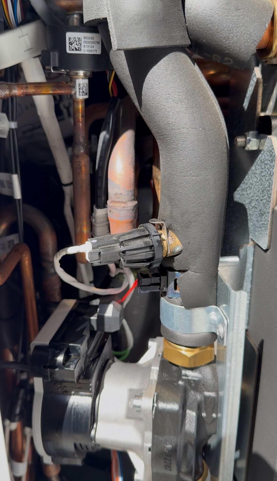 Fragment instalacji z rurami miedzianymi, czarną izolacją termiczną, czujnikiem i elementami mocującymi. Widoczne detale przyłączeń i przewodów w kotłowni.