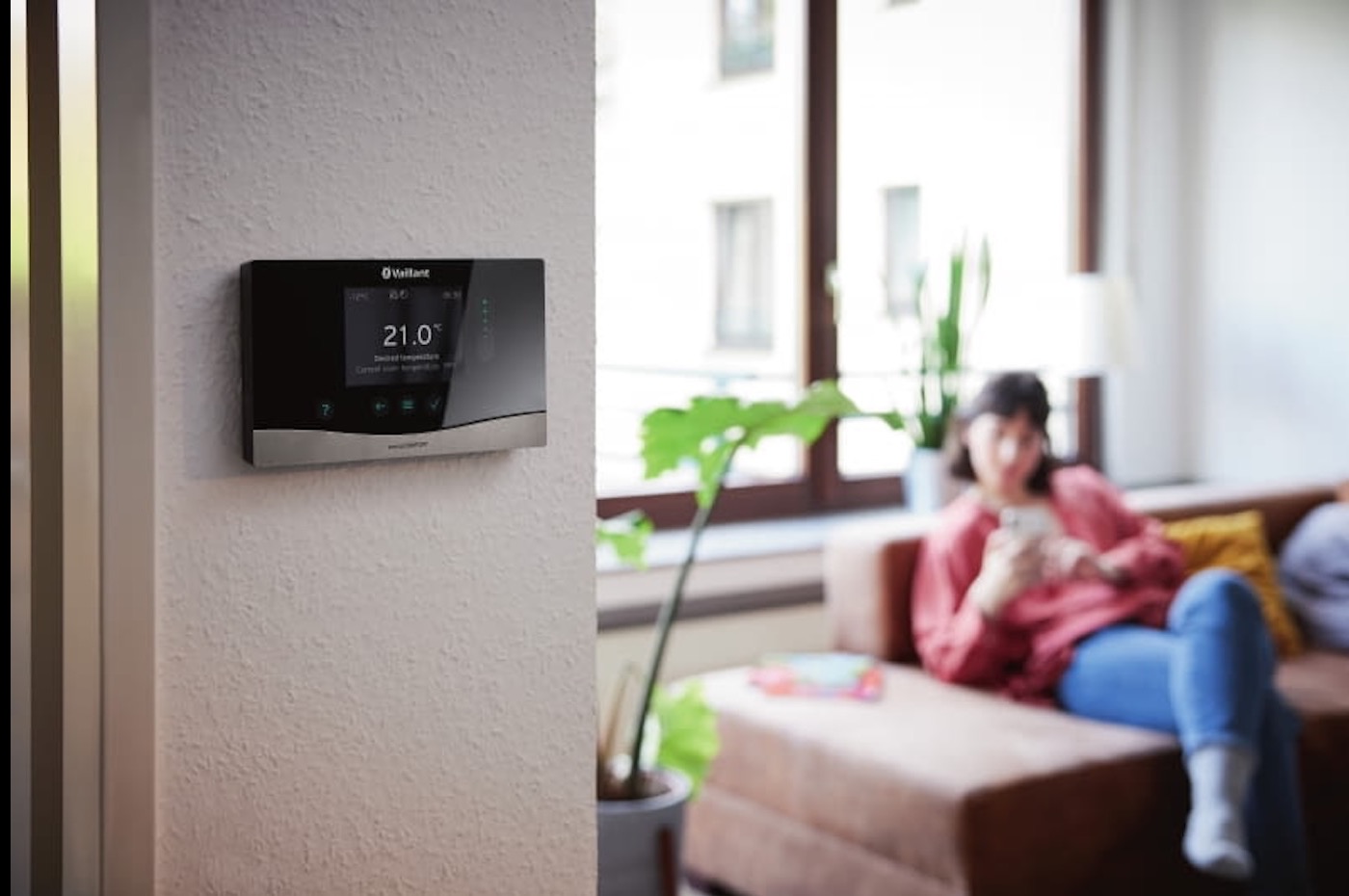 Nowoczesny panel sterowania ogrzewaniem Vaillant na ścianie w salonie, w tle kobieta na kanapie. Wyświetlacz pokazuje 21 stopni Celsjusza.