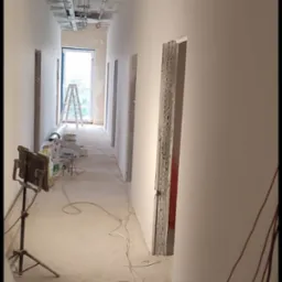 Korytarz w trakcie prac wykończeniowych: świeżo pomalowane ściany, instalacje pod sufitem, otwory drzwiowe, drabina w tle. Widoczne narzędzia i kable na podłodze.