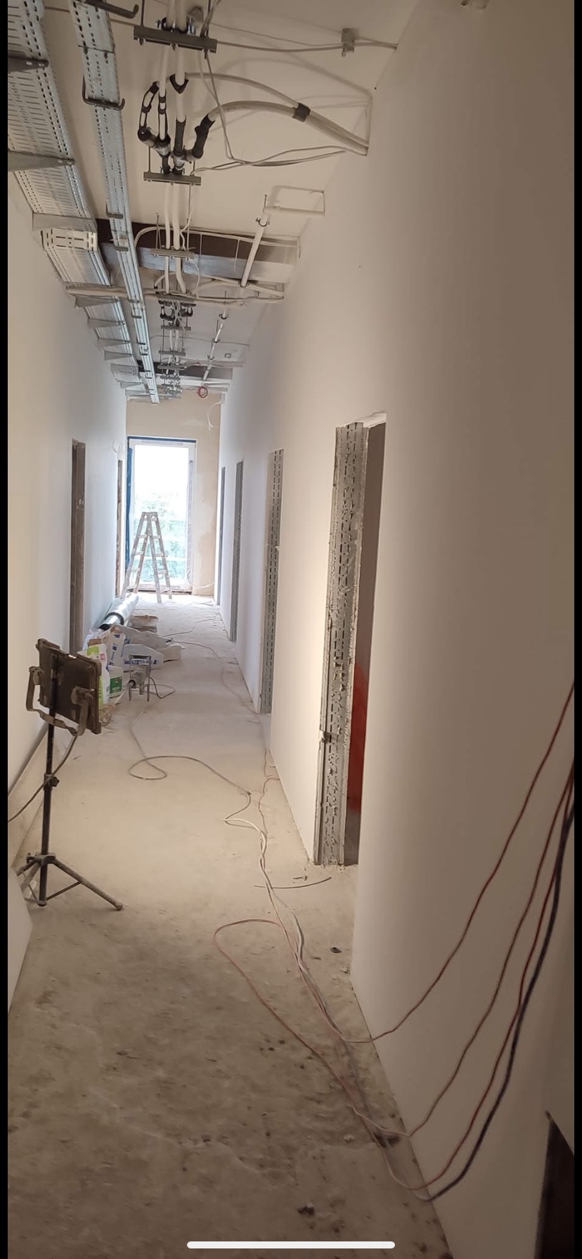 Korytarz w trakcie prac wykończeniowych: świeżo pomalowane ściany, instalacje pod sufitem, otwory drzwiowe, drabina w tle. Widoczne narzędzia i kable na podłodze.