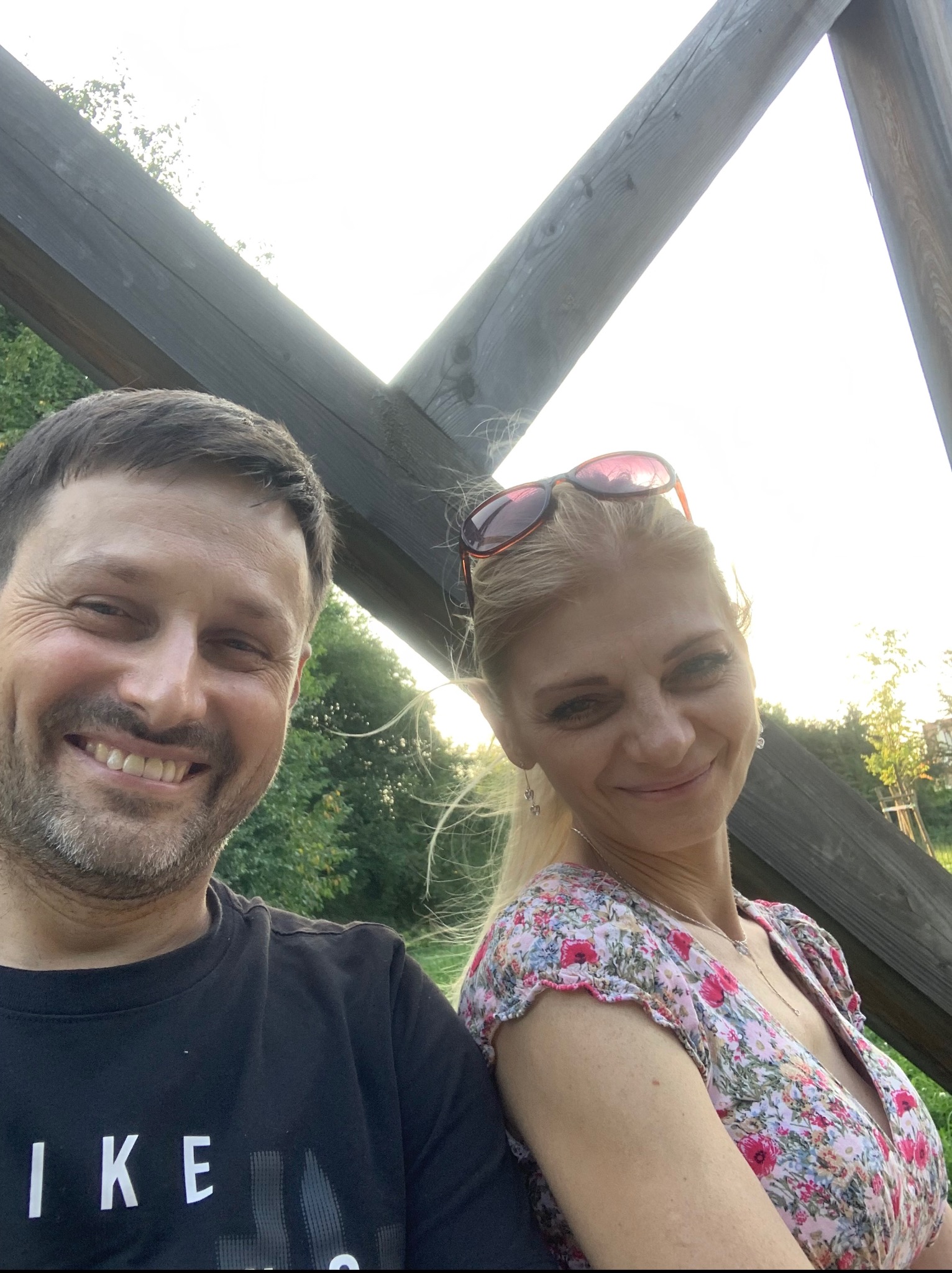 Uśmiechnięta para w Krakowie siedzi na drewnianej konstrukcji, w tle zieleń. Kobieta w kwiecistej sukience i okulary na głowie, mężczyzna w czarnym t-shircie.