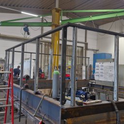 WELT Paweł Brak - Wnętrze warsztatu z konstrukcją z metalowych profili w trakcie budowy. Widoczny spawacz i narzędzia. Elementy konstrukcyjne w surowym stanie, bez wykończenia.