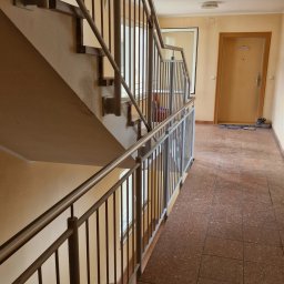 WELT Paweł Brak - Klatka schodowa w bloku z metalowymi, pionowymi barierkami. Widok z dołu na schody i poręcze. Jasne ściany i brązowa podłoga z płytek.