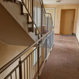 Klatka schodowa w bloku z metalowymi, pionowymi barierkami. Widok z dołu na schody i poręcze. Jasne ściany i brązowa podłoga z płytek.