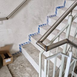 WELT Paweł Brak - Metalowe poręcze i balustrady na betonowych schodach w trakcie montażu, widoczne zabezpieczenia na stopniach. Karton na podłodze. Ujęcie z góry.