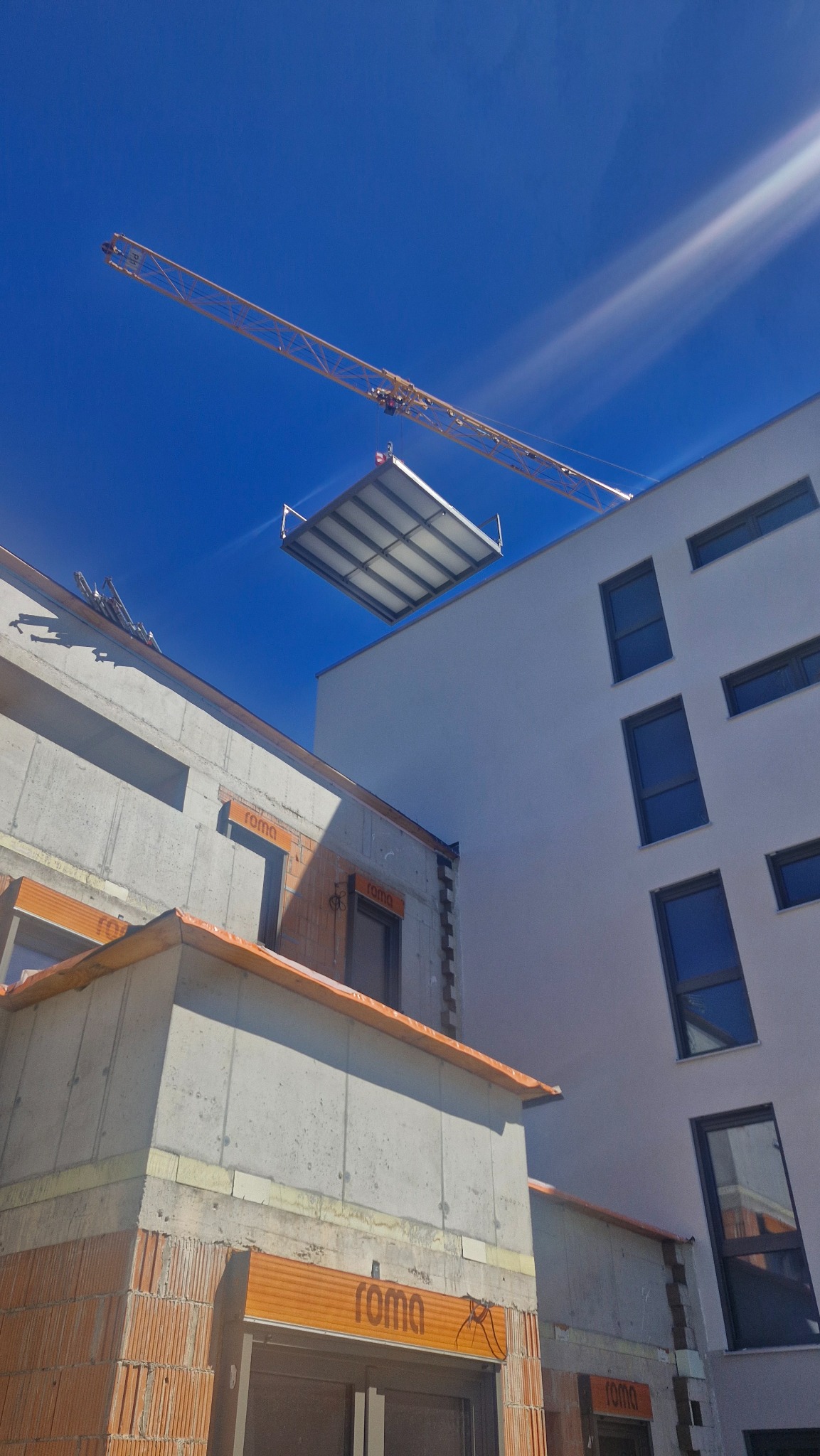 Żuraw unosi element konstrukcyjny na tle budynków w trakcie budowy, błękitne niebo w tle. Element konstrukcyjny z metalową ramą.